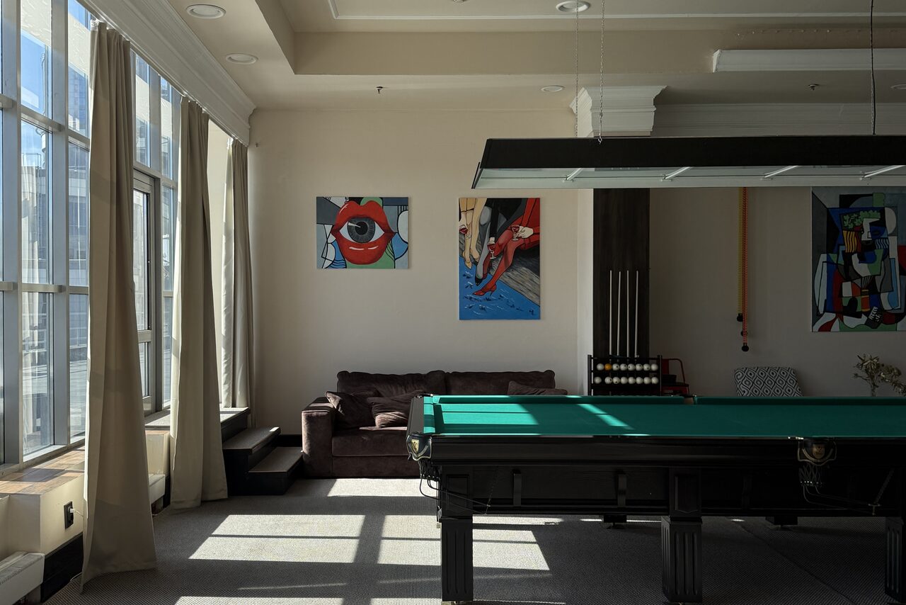 Арендовать Студия Н. Пространство с биллиардным столом, коричневым диваном, большими окнами с занавесками. На стенах три яркие картины. Над столом висят светильники. Светло-серые стены и пол, деревянные элементы интерьера.