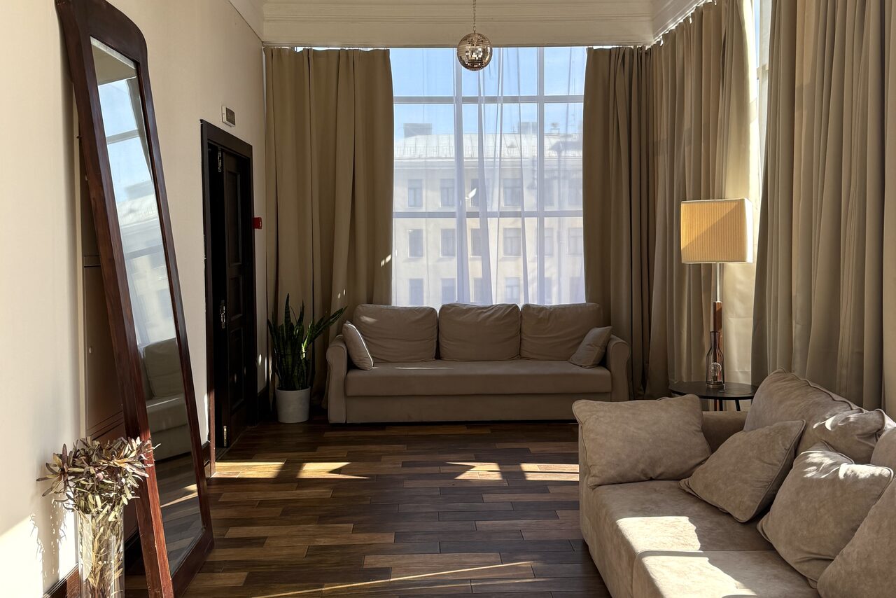 Арендовать Студия Н. Светлая гостиная с двумя бежевыми диванами, большим зеркалом в темной раме, напольной лампой и комнатным растением. Паркетный пол, окна с занавесками, здание на улице виднеется через стекло.