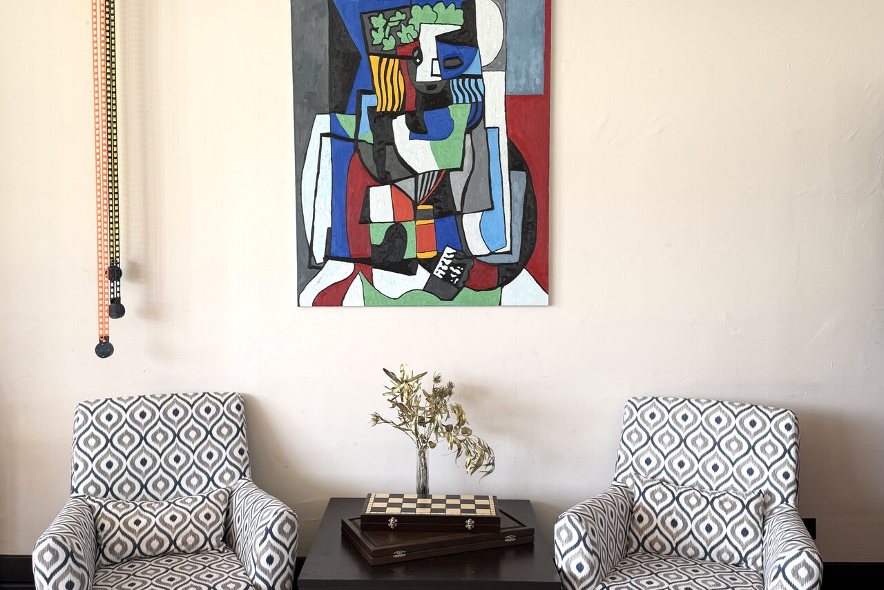 Арендовать Студия Н. Две узорчатые кресла возле квадратного стола с настольной игрой и сухими цветами. На светлой стене висит абстрактная картина. Вертикальные светильники слева. Светлый интерьер с геометрическими и динамичными элементами.