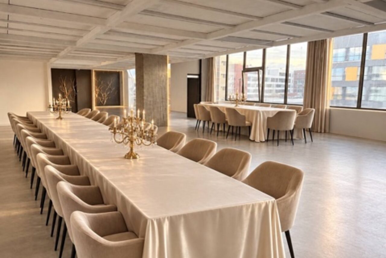Арендовать Loft Prostranstvo Huge(Огромное Лофт Пространство). Просторное помещение с большими окнами, длинный стол с белой скатертью и золотыми канделябрами, мягкие стулья бежевого цвета, пол из светлого дерева или ламината, потолок с балками, светлые стены, много естественного света.