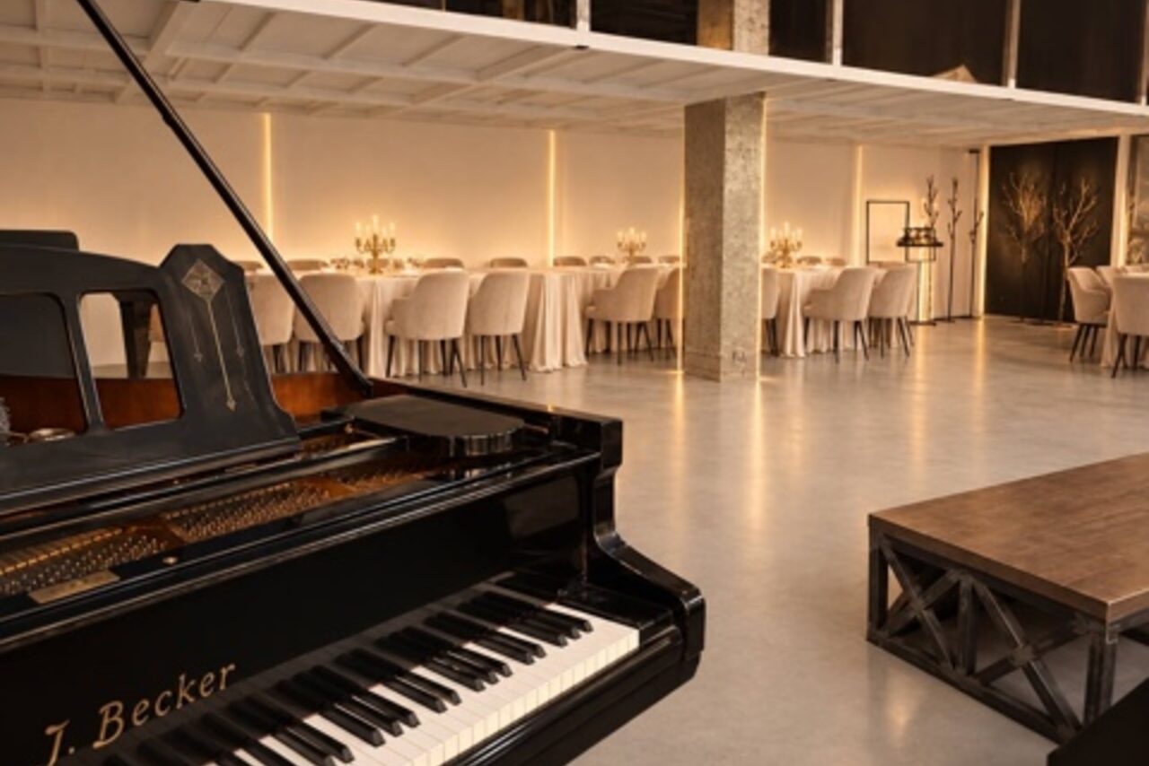 Арендовать Loft Prostranstvo Huge(Огромное Лофт Пространство). Помещение со столами и стульями, украшенными скатертями и чехлами, в центре черный рояль, справа деревянный стол. Стены светлые с мягкой подсветкой. Пол бетонный. Открытая планировка, высокие потолки.