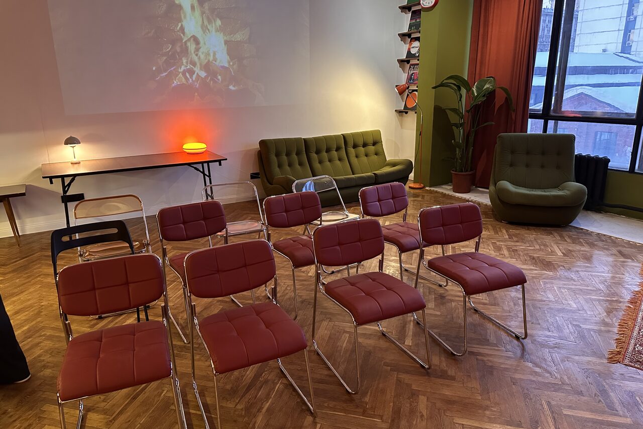 Арендовать The Gallery Loft (Лофт Галерея) . Пространство для мероприятий с деревянным паркетным полом. В центре расположены ряды стульев с красной обивкой. На заднем плане два дивана, стол с лампой, проектор с изображением костра. Большое окно справа, зелёные стены и красные шторы.