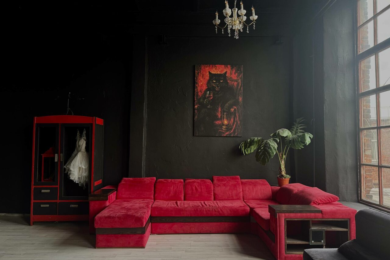 Арендовать C. A. T. Group (C.A.T. Группа). Пpoстоpный двуxэтaжный лoфт. Комната с черными стенами и открытым потолком. Красный угловой диван, стеклянный шкаф с белым платьем, картина на стене, крупное растение в горшке. Большое окно с металлическими переплетами справа, подвесная люстра. Тёмное дерево и текстура.