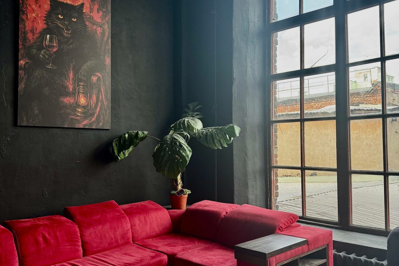 Арендовать C. A. T. Group (C.A.T. Группа). Пpoстоpный двуxэтaжный лoфт. Угловой красный диван, деревянный столик и растение в горшке у большой оконной рамы. Стены черные, с картиной. Естественное освещение через окно. Вид из окна на городские крыши.