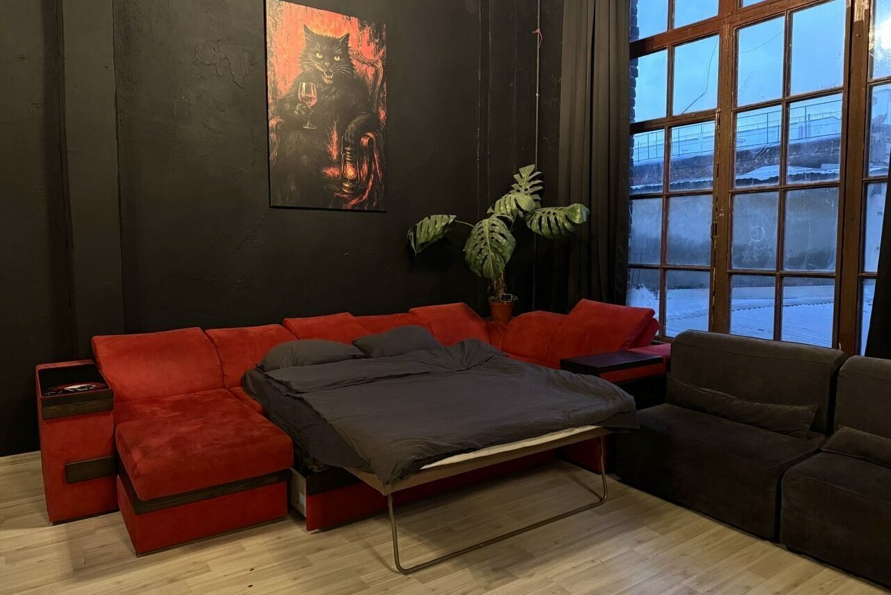 Арендовать C. A. T. Group (C.A.T. Группа). Пpoстоpный двуxэтaжный лoфт. Комната с красным угловым диваном-кроватью и двумя черными креслами. Темные стены, картина с изображением животного, крупное окно с темными шторами, напольное покрытие светлое, большое растение в горшке рядом с диваном.