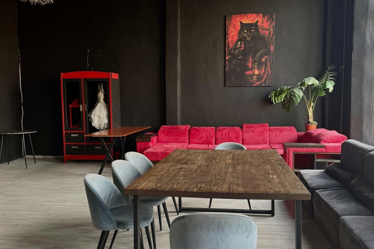Арендовать C. A. T. Group (C.A.T. Группа). Пpoстоpный двуxэтaжный лoфт. Просторное помещение с черными стенами, деревянным полом. Красный диван, серый стул, деревянный стол. Красная гардеробная с белым платьем. На стене картина с изображением кошки. Растение в горшке, большое окно справа.