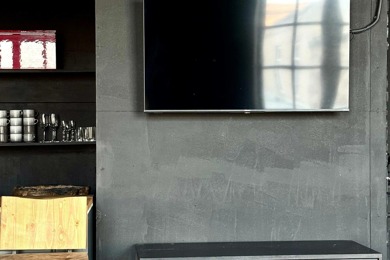 Арендовать C. A. T. Group (C.A.T. Группа). Пpoстоpный двуxэтaжный лoфт. Стена с серым покрытием, на ней висит большой телевизор. Слева черные полки с чашками и бокалами. Внизу видна деревянная деталь. Освещение естественное, с окна отражаются блики на телевизоре.