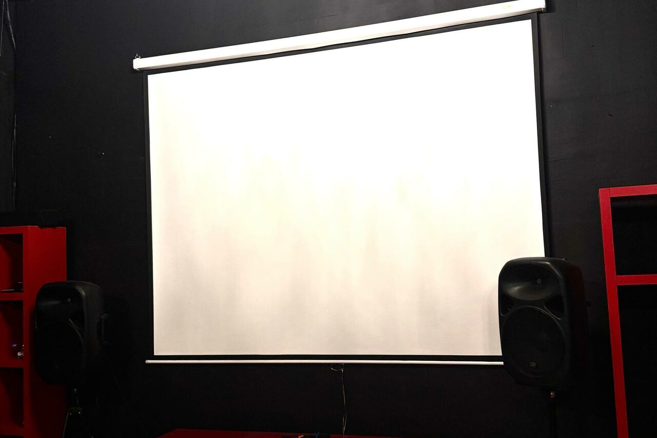 Арендовать C. A. T. Group (C.A.T. Группа). Пpoстоpный двуxэтaжный лoфт. Экран для проектора на черной стене, по бокам две акустические колонки. Красные стеллажи видны частично. Освещение отсутствует. Общий вид минимум мебели и оборудования.