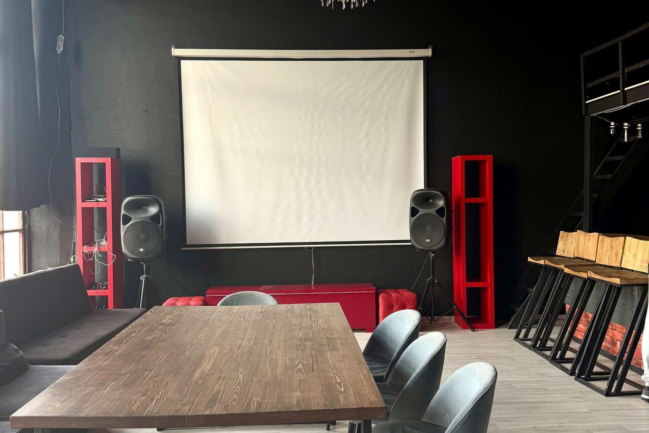 Арендовать C. A. T. Group (C.A.T. Группа). Пpoстоpный двуxэтaжный лoфт. Черное помещение с экраном для проектора, два динамика, деревянный стол, серые кресла, черные стулья у высокой стойки, красные декоративные элементы, деревянный пол. Стеллажи и оборудование размещены по периметру. Слабое естественное освещение.