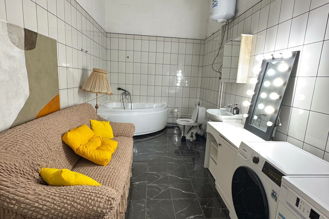 Арендовать C. A. T. Group (C.A.T. Группа). Пpoстоpный двуxэтaжный лoфт. Комната с кафельными стенами и черным мраморным полом. Угловая ванна, рядом унитаз и раковина. На стене большое зеркало с лампами. Плетеный диван с желтыми подушками, настольная лампа. Стиральная и сушильная машины. Абстрактная картина на стене.