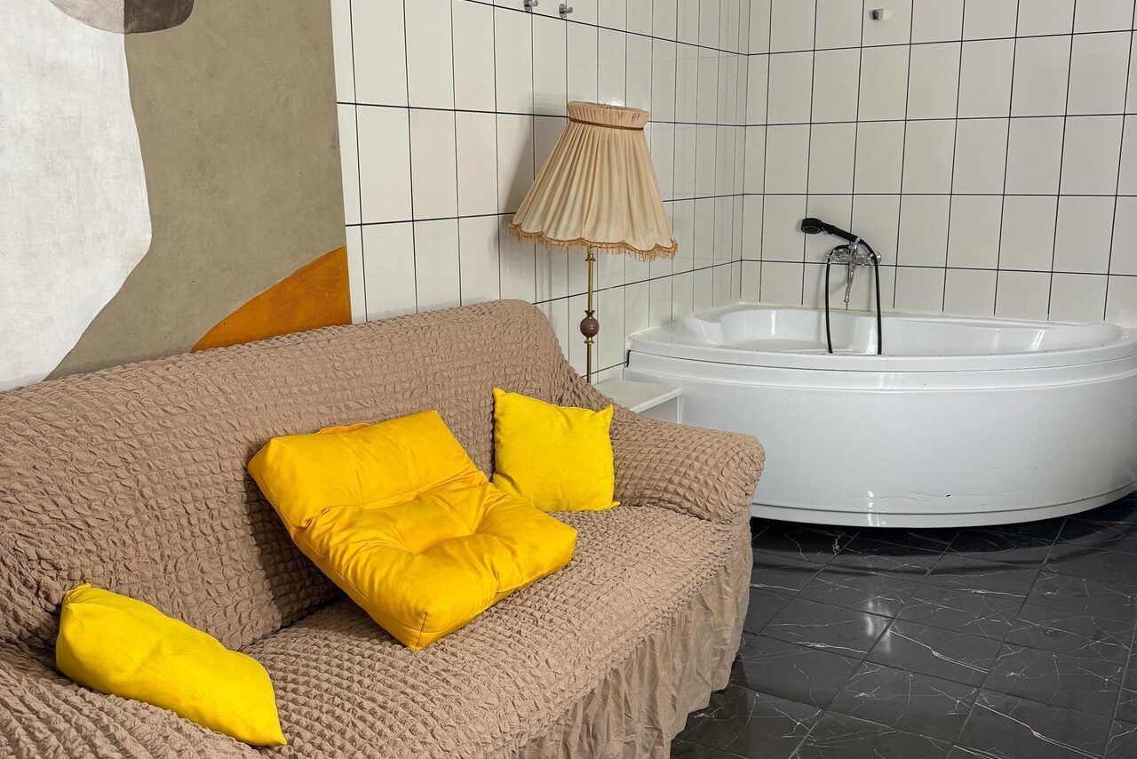 Арендовать C. A. T. Group (C.A.T. Группа). Пpoстоpный двуxэтaжный лoфт. Меблированное пространство с коричневым диваном с жёлтыми подушками, белая угловая ванна, напольная лампа с бежевым абажуром. Стены облицованы белой плиткой, пол из тёмного камня или плитки. Видна часть стены с абстрактным рисунком.