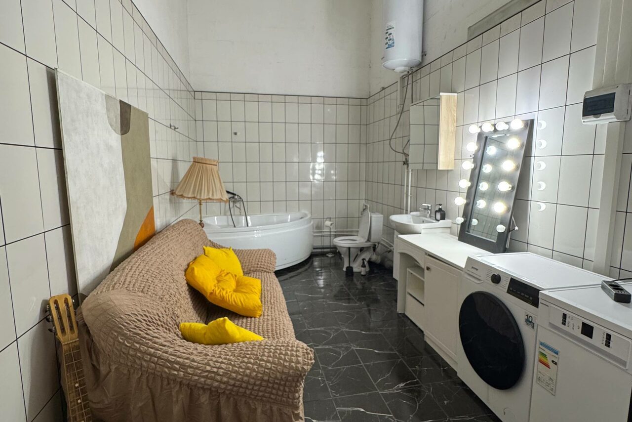 Арендовать C. A. T. Group (C.A.T. Группа). Пpoстоpный двуxэтaжный лoфт. Пространство с ванной и стиральной машиной, угловая планировка. Диван с бежевым покрывалом и жёлтыми подушками, круглое зеркало с лампочками, кафельные стены, чёрный пол. Ванная, унитаз, водонагреватель. Лампа с абажуром, декоративная картина.