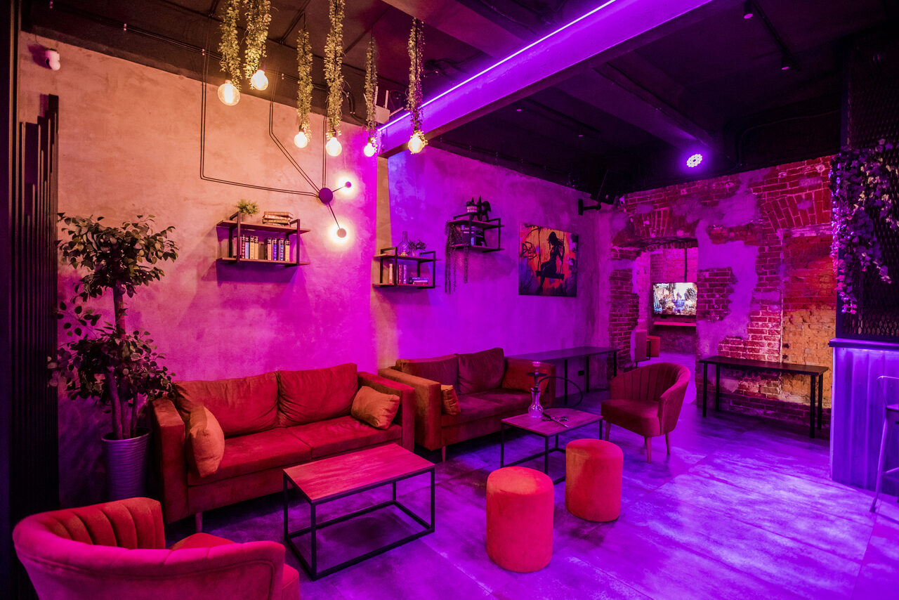 Арендовать Дизайнерский лофт с караоке и активностями возле метро. Лофт-пространство с кирпичными стенами, диванами и креслами красного цвета. На потолке висят лампы накаливания. Есть растения и полки с книгами. Освещение фиолетовое. На стене картина и висят декоративные элементы. В комнате стулья и журнальные столики.