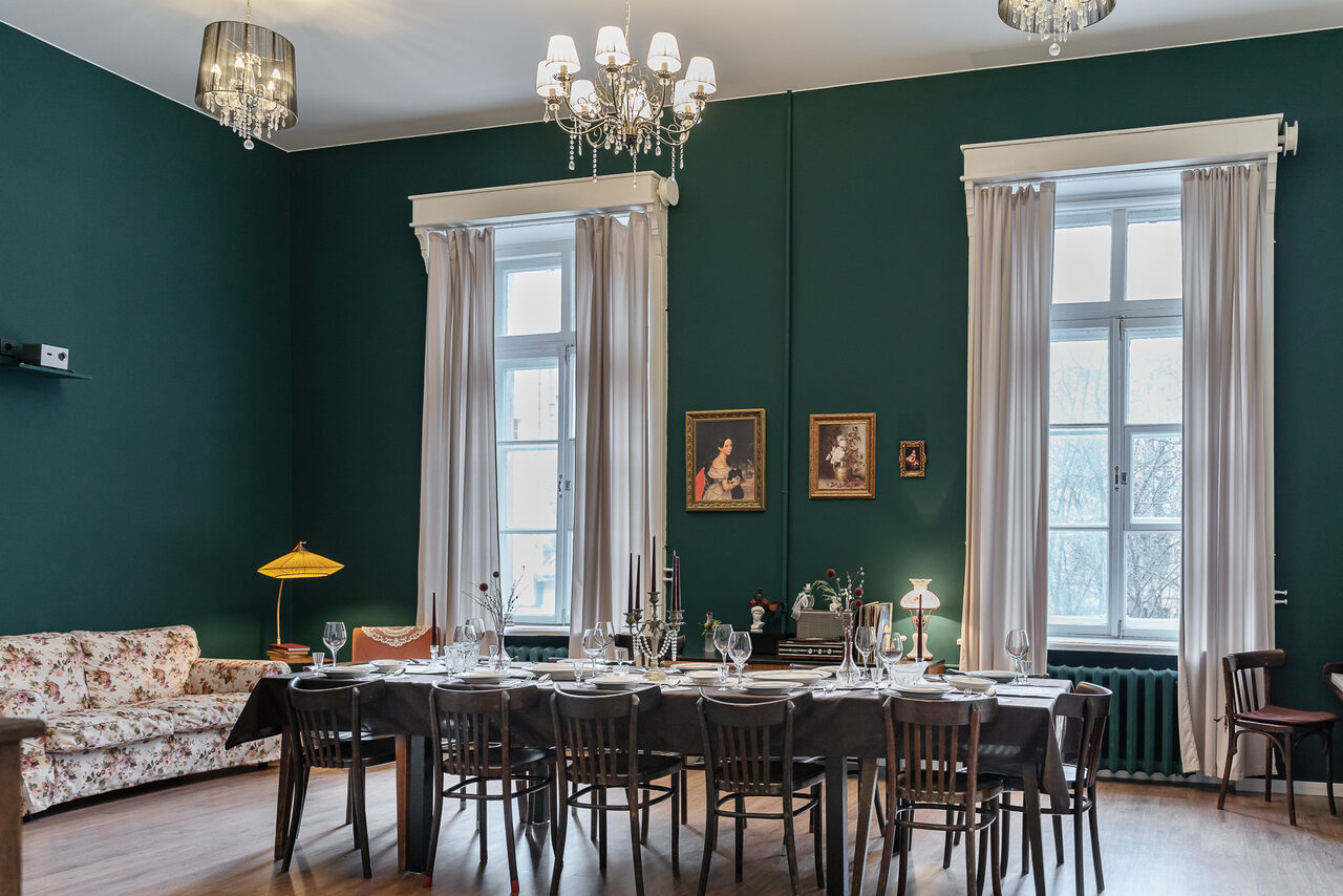 Арендовать Пространство Имена. Винтажный лофт . Просторная комната с тёмно-зелёными стенами и белым потолком. Большой стол, сервированный для ужина, окружён деревянными стульями. Справа — две высокие окна с белыми шторами. На стене — картины. Люстра и настольные лампы обеспечивают освещение.