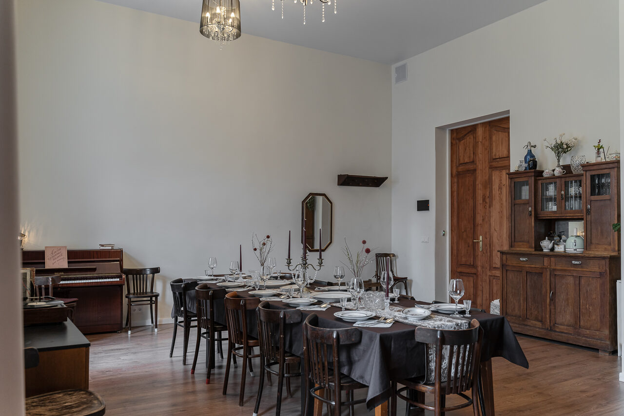 Арендовать Пространство Имена. Винтажный лофт . Большая комната с деревянным полом, длинным столом, накрытым темной скатертью, вокруг деревянные стулья. Справа деревянный шкаф, слева пианино. На столе посуда и бокалы. На стенах светлые обои, в центре подвесной светильник.