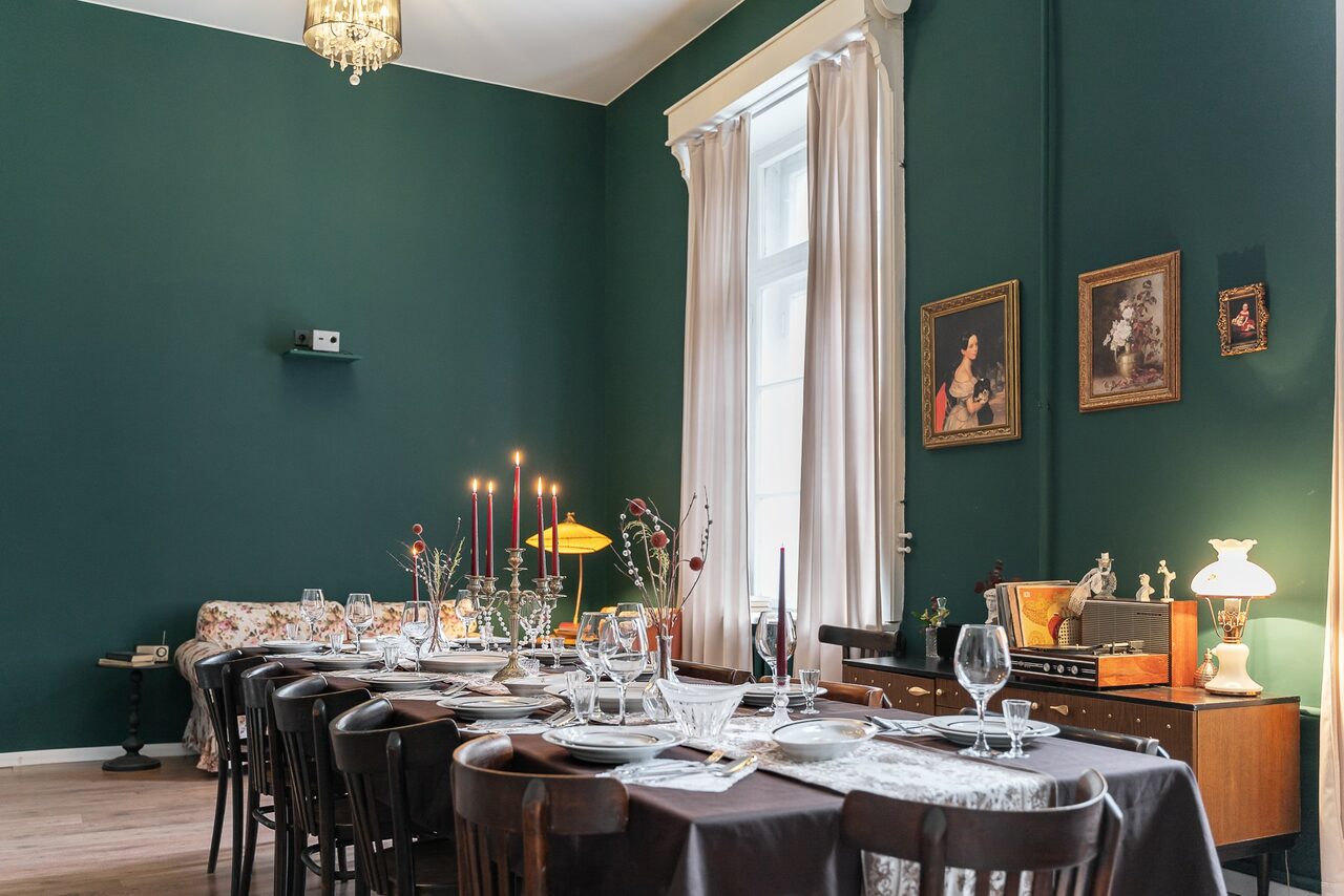 Арендовать Пространство Имена. Винтажный лофт . Столовая с зеленым интерьером, длинным столом и черными стульями. На столе белая посуда и бокалы. На заднем плане диван, комод с лампами и картинами. Высокие окна с белыми занавесками. Светильник на потолке.