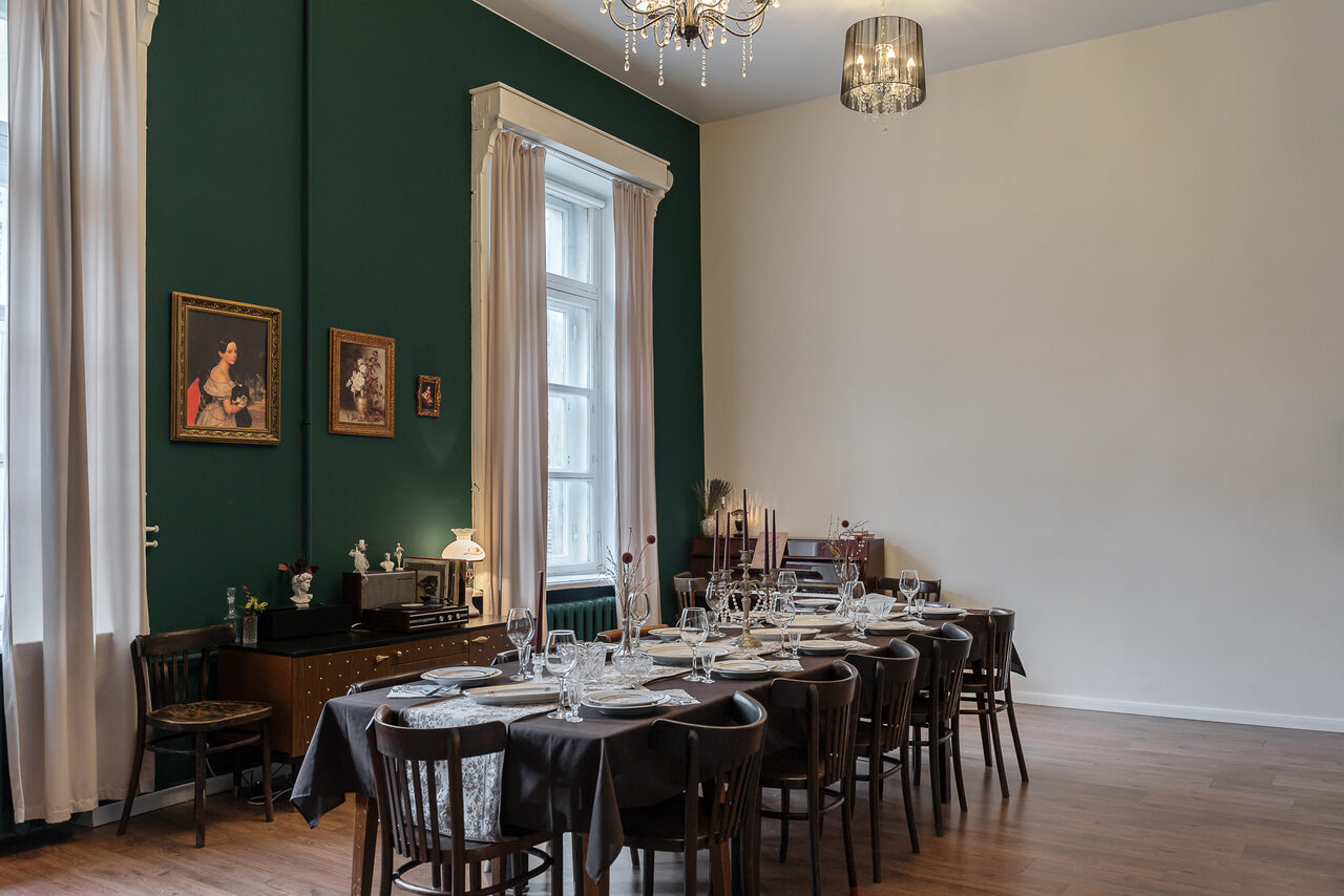 Арендовать Пространство Имена. Винтажный лофт . Комната с длинным обеденным столом, сервированным посудой и бокалами. Стулья деревянные, тёмные. Стены зелёного и белого цвета, на зелёной стене картины. Деревянный пол, большие окна с белыми шторами. Классические светильники, полки с украшениями.