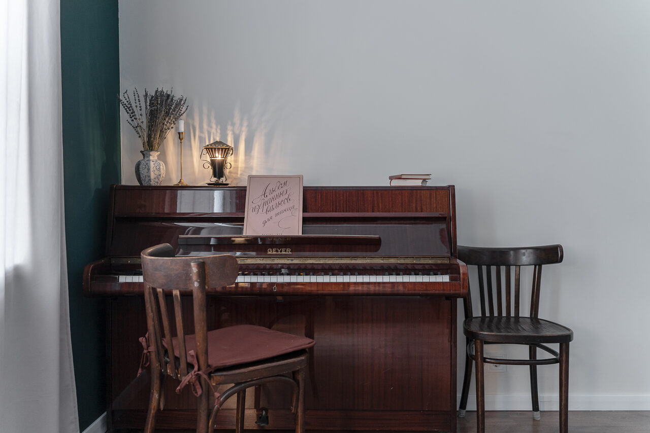 Арендовать Пространство Имена. Винтажный лофт . Комната с вертикальным пианино, двумя деревянными стульями, вазой с сухими цветами, настольной лампой и книгами на пианино. Стены белые и зелёные, пол тёмный. Освещение мягкое, мебель деревянная.