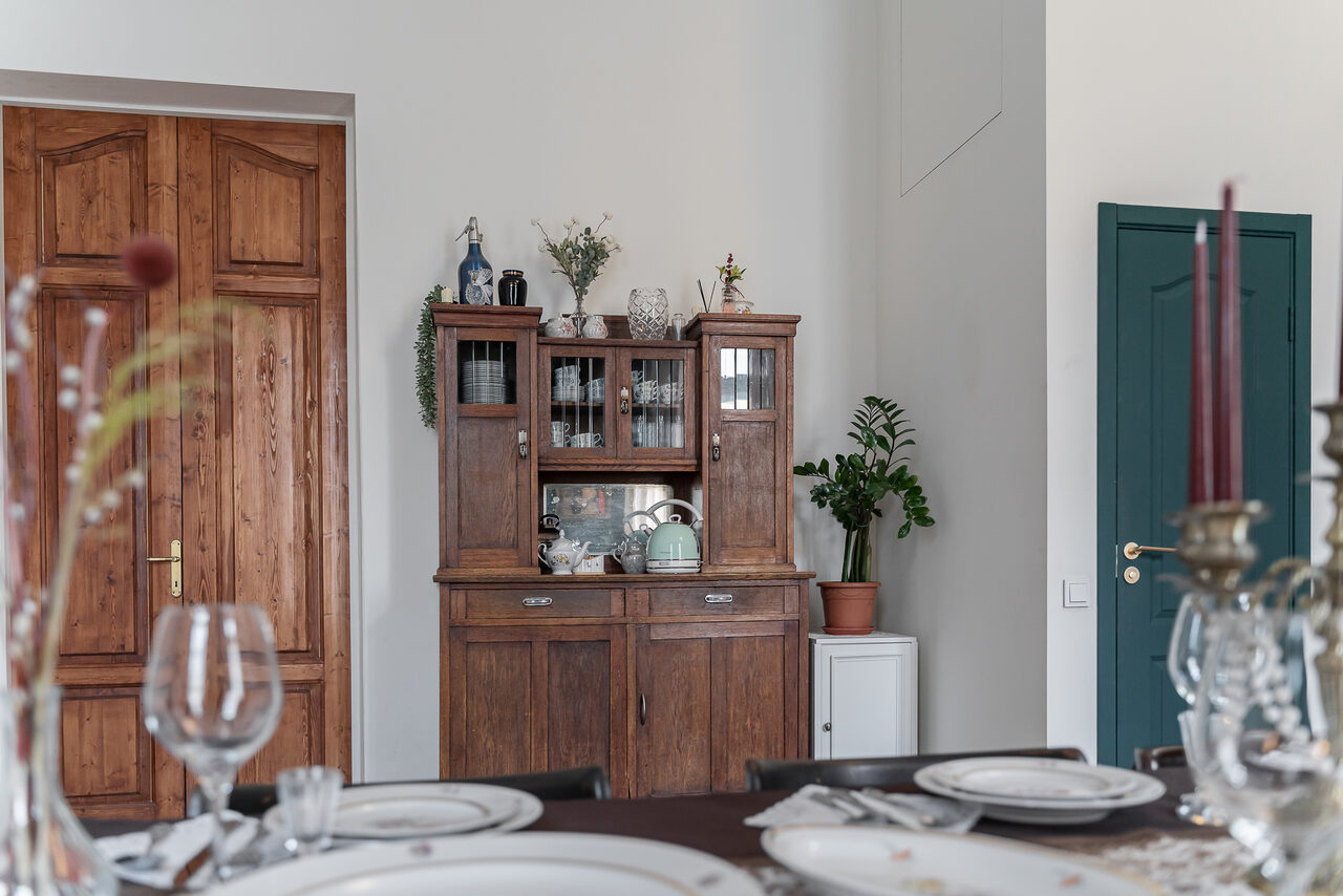 Арендовать Пространство Имена. Винтажный лофт . Столовая комната с деревянным буфетом, обставленным посудой и декоративными элементами. На переднем плане накрытый обеденный стол с тарелками и бокалами. Светлые стены, деревянные двери, зелёное растение в углу.
