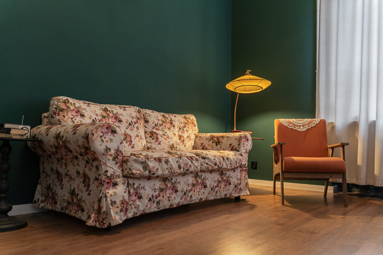 Арендовать Пространство Имена. Винтажный лофт . Гостиная с зелеными стенами, на полу деревянное покрытие. Диван в цветочной обивке, рядом оранжевое кресло. Торшер с абажуром, белые шторы на окне. Небольшой стол с книгой и лампой.