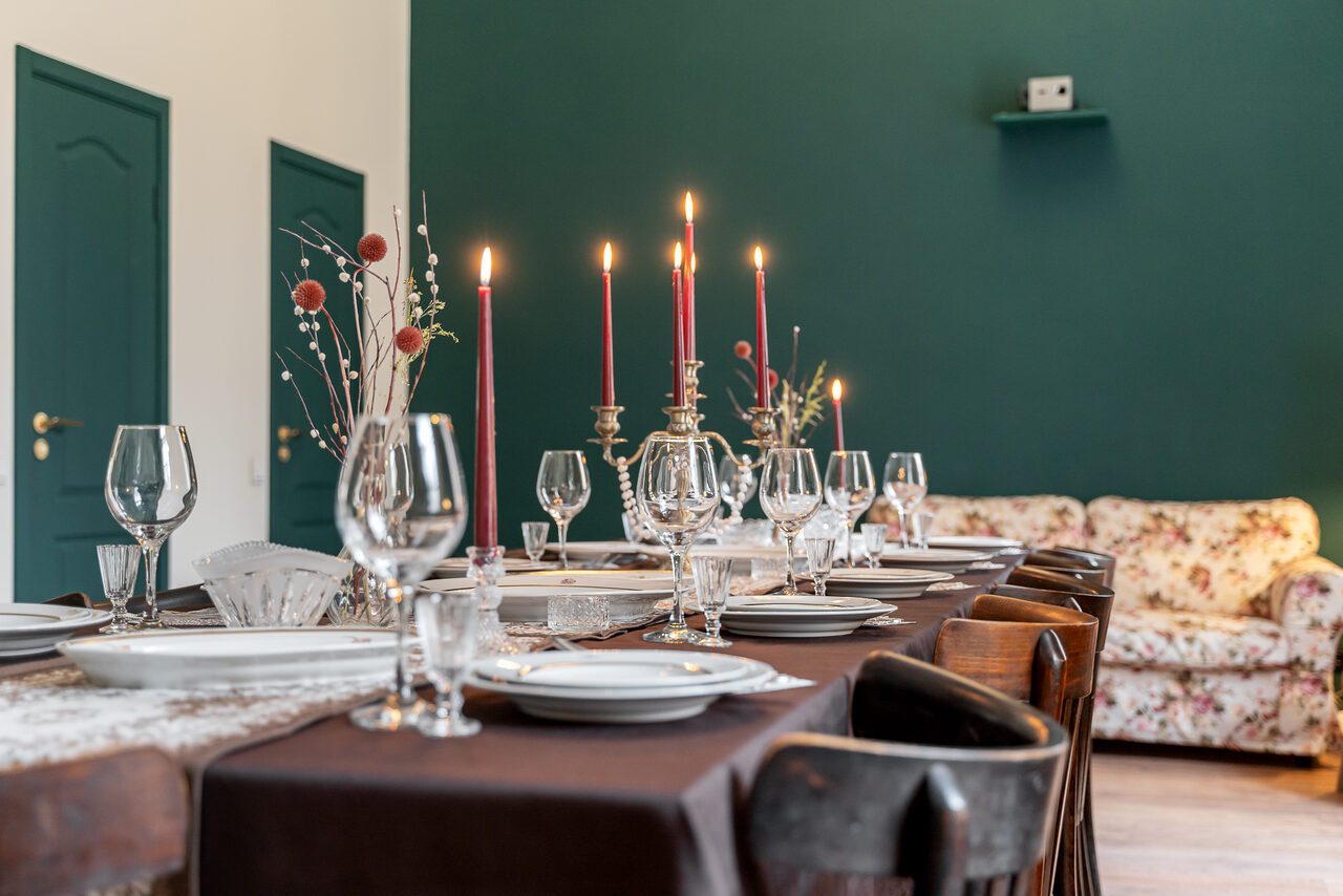 Арендовать Пространство Имена. Винтажный лофт . Столовая с длинным деревянным столом, накрытым темной скатертью. На столе расставлены свечи в канделябрах, бокалы, тарелки и декор. На заднем плане диван с цветочным орнаментом, зеленая стена с дверями, пол деревянный.