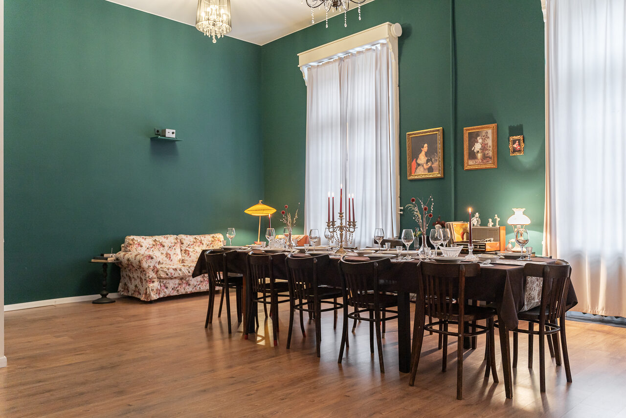 Арендовать Пространство Имена. Винтажный лофт . Комната с высокими потолками, стены зеленого цвета. В центре длинный деревянный стол с накрытой посудой, окруженный стульями. Справа диван с цветочным узором. На стене несколько картин. Большие окна с длинными шторами. Деревянный пол.