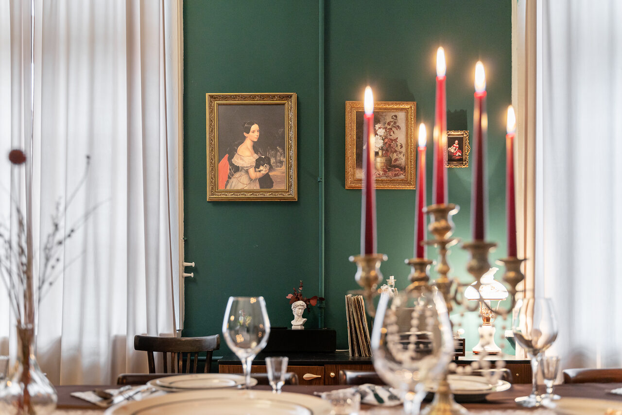 Арендовать Пространство Имена. Винтажный лофт . Столовая с зелёными стенами и белыми шторами. На стене картины в рамах. Большой стол с белой посудой и бокалами. На столе высокий подсвечник с зажжёнными свечами. Деревянные стулья. Тёплое освещение.
