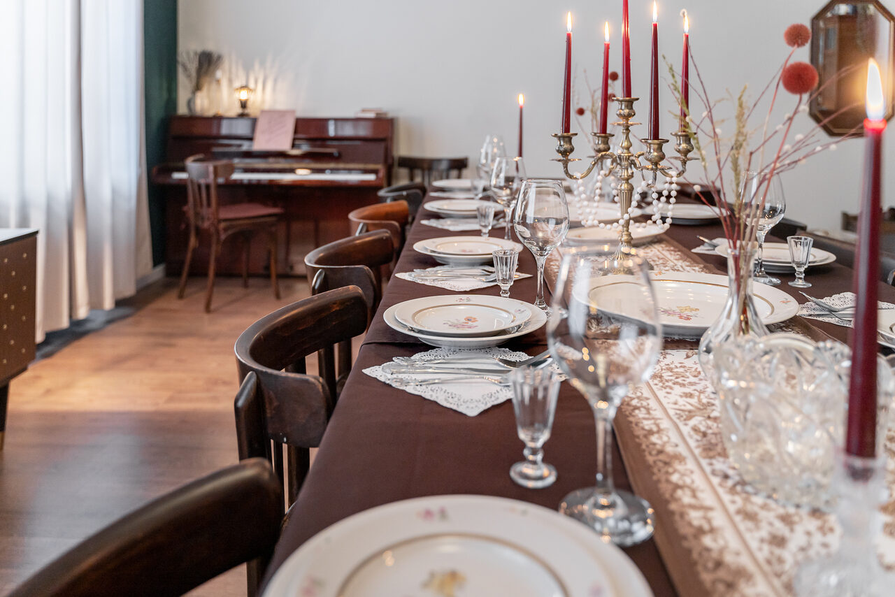 Арендовать Пространство Имена. Винтажный лофт . Обеденная зона с длинным столом, покрытым темной скатертью. На столе белая посуда, стеклянные бокалы, красные свечи в латунных канделябрах. Кресла из темного дерева. На фоне фортепиано и окна с белыми шторами. Светлая деревянная полка.