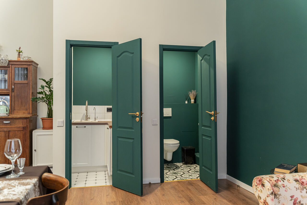 Арендовать Пространство Имена. Винтажный лофт . Кухонная зона с деревянными и зелеными элементами; две двери, за одной из которых ванная комната с узорчатым полом и унитазом, за другой — раковина. Стол с тарелками и бокалами. Потолки высокие, стены зелёные.