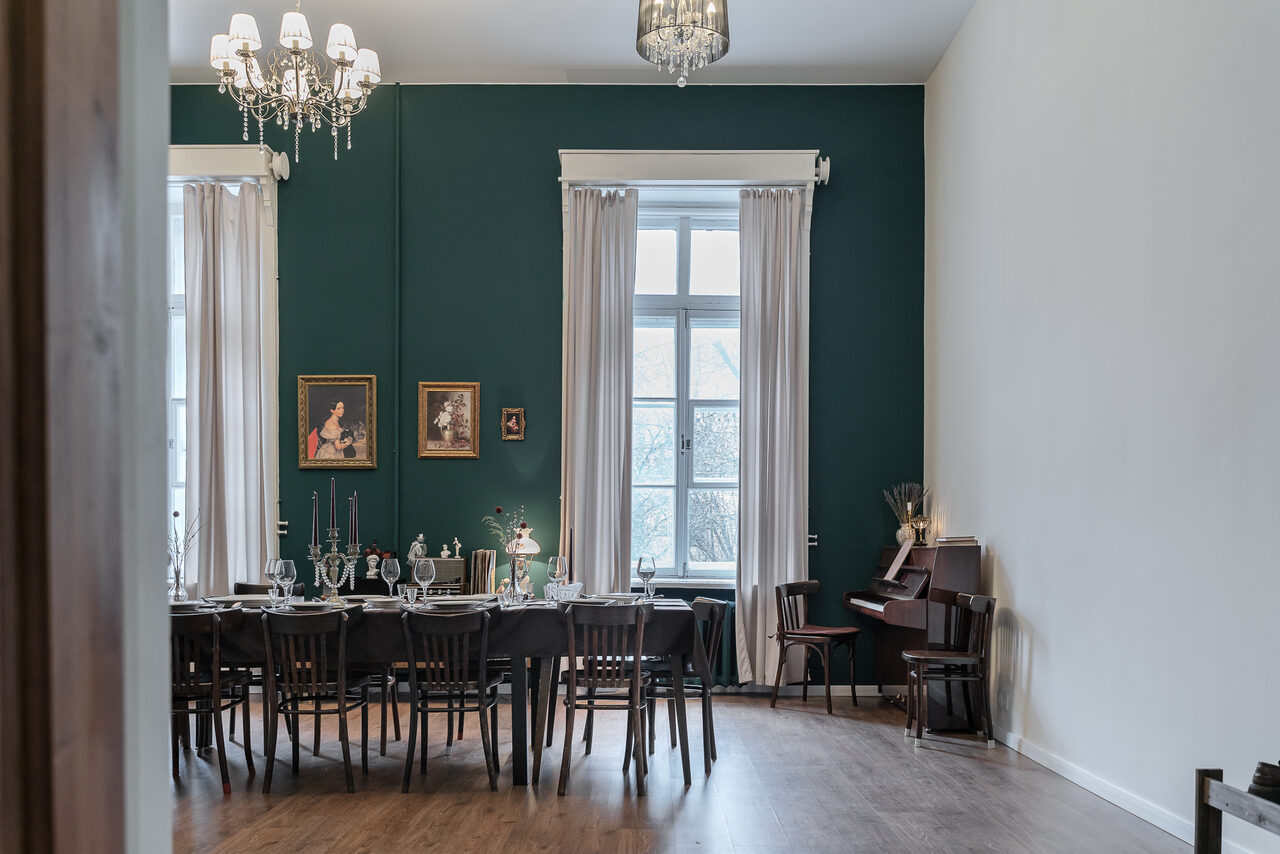 Арендовать Пространство Имена. Винтажный лофт . Просторная комната с высокими потолками и деревянным полом. Стол с черной скатертью и стульями вдоль зеленой стены. Пианино справа, над столом висят люстры. Белые шторы на больших окнах. На стене картины в рамках.