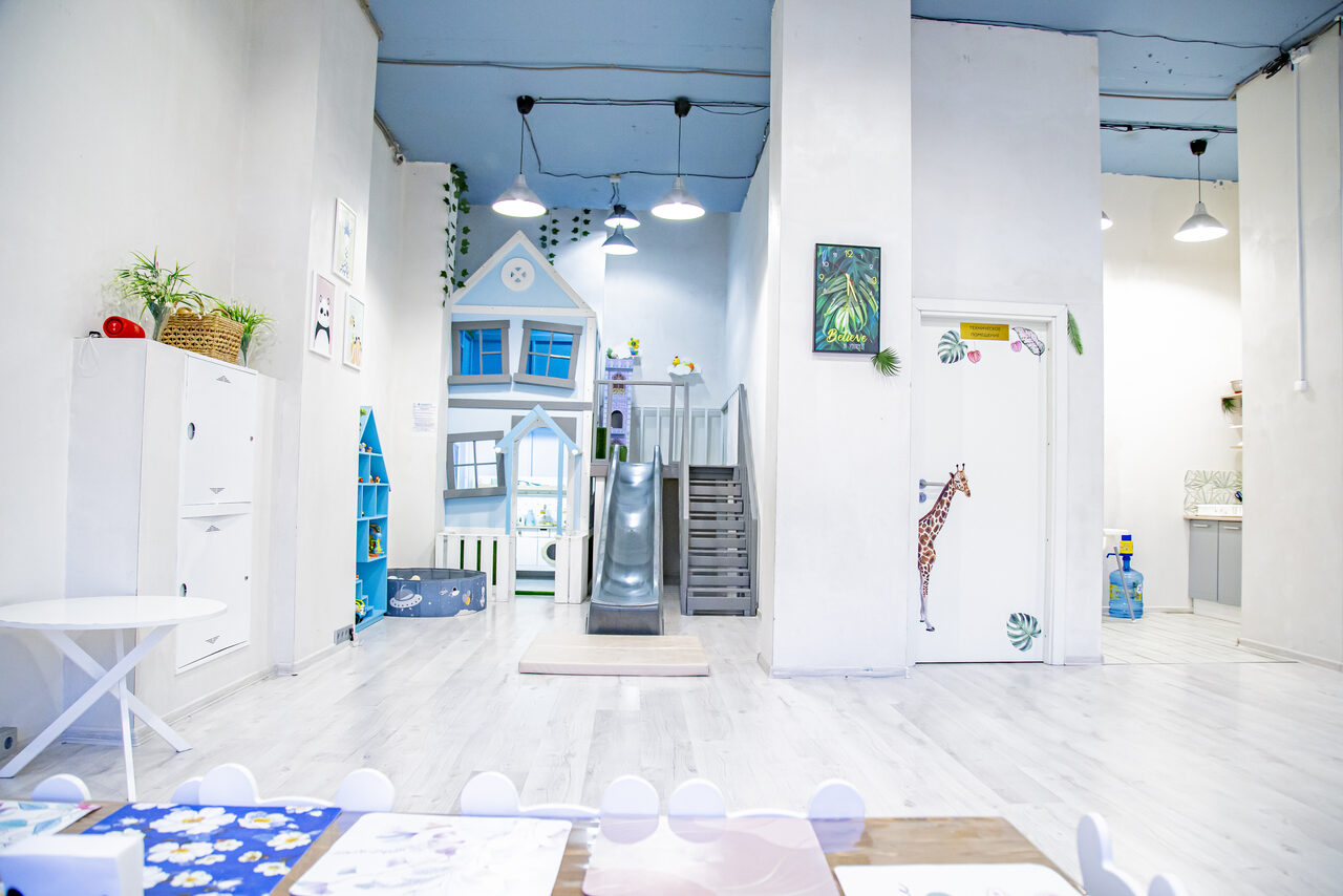 Арендовать Лофт Облака. Зал Баунти на Полярной. Детская игровая комната с деревянными полами и белыми стенами, оформленная в светлых цветах. Возвышение с лесенкой и горкой, декоративный домик, наклейка с жирафом на двери. Стол с белыми стульями и цветной скатертью, зелёные растения на полках.