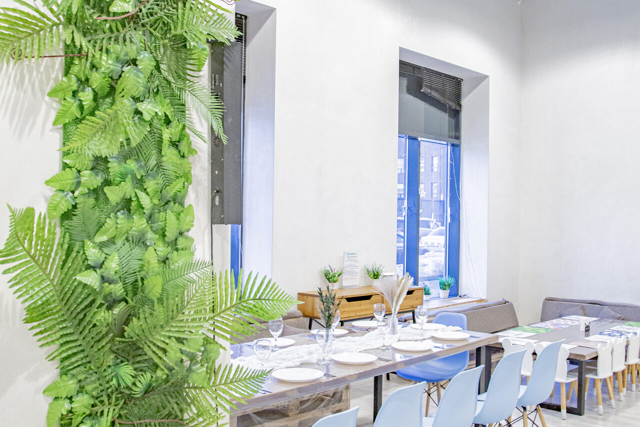 Арендовать Лофт Облака. Зал Баунти на Полярной. Просторное светлое помещение с длинным столом, сервированным тарелками и бокалами, окруженным голубыми стульями. На стене зеленая растительность. Два больших окна с видом на улицу. Нейтральные стены, натуральное и искусственное освещение.