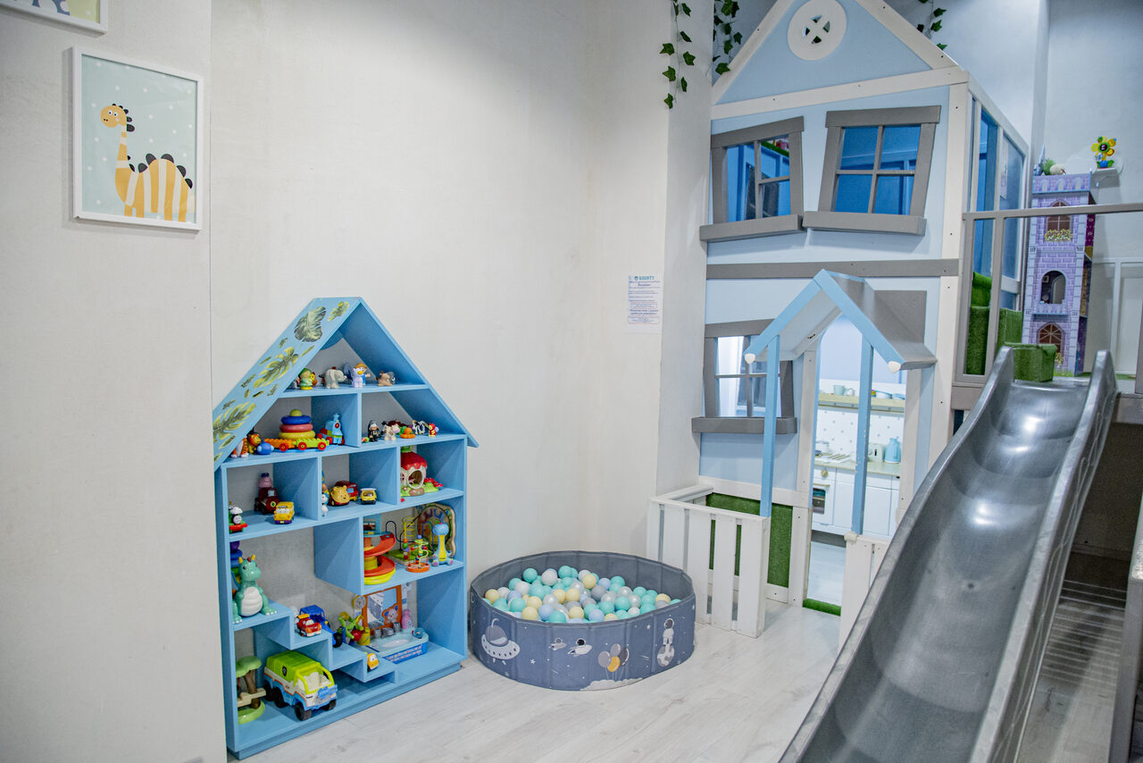 Арендовать Лофт Облака. Зал Баунти на Полярной. Детская комната с горкой, домиком с окнами, игрушечной мебелью, синими стеллажами с игрушками и пластиковым бассейном с разноцветными шарами. Белый пол, светлые стены, теплое освещение. Картина с изображением жирафа на стене.