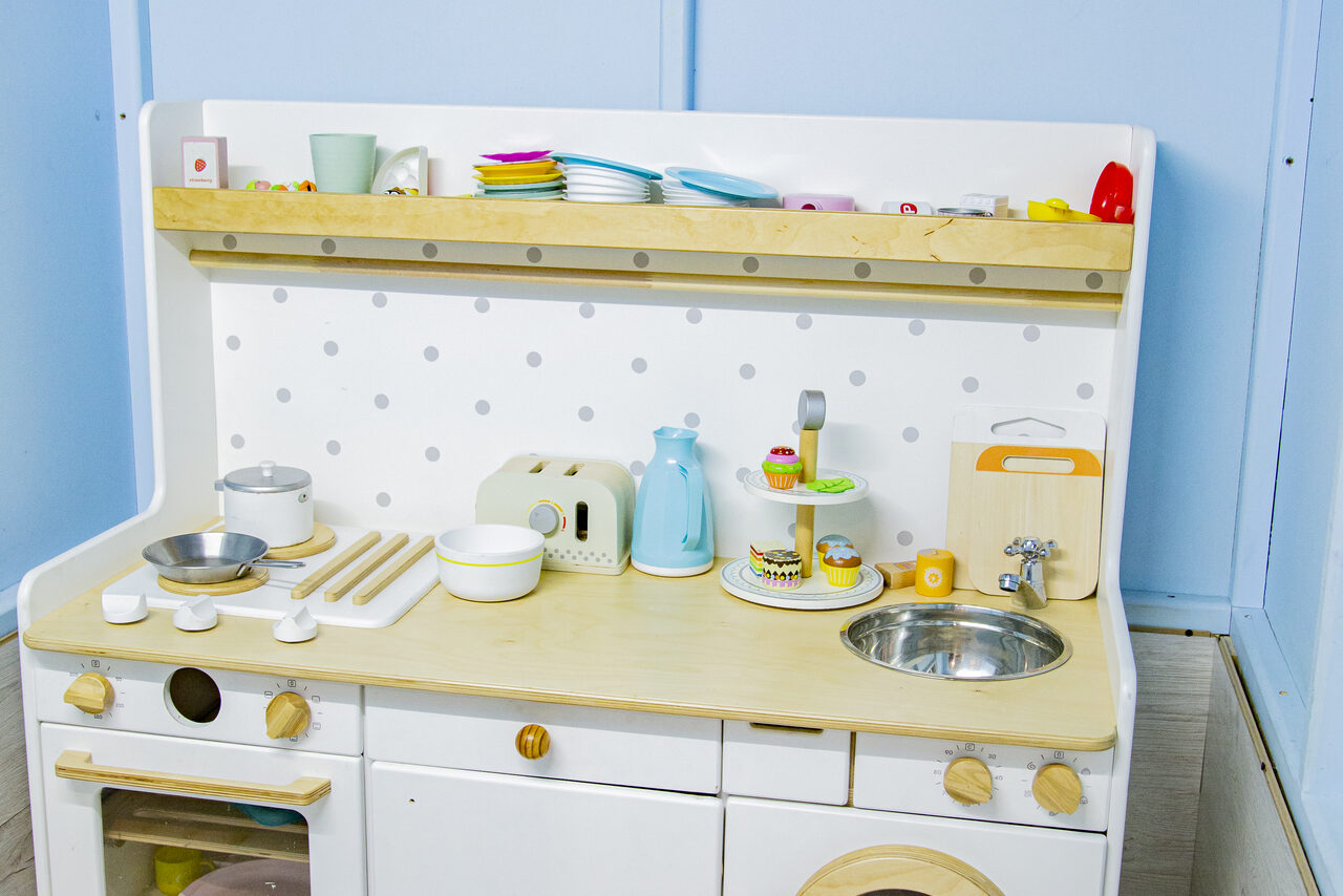 Арендовать Лофт Облака. Зал Баунти на Полярной. Детская игровая кухня с деревянной плоской столешницей и белыми элементами. Полка с разноцветной детской посудой над столешницей. Имитация раковины и плиты, игрушечные приборы, кухонные принадлежности. Стена с голубыми и серыми полосами.