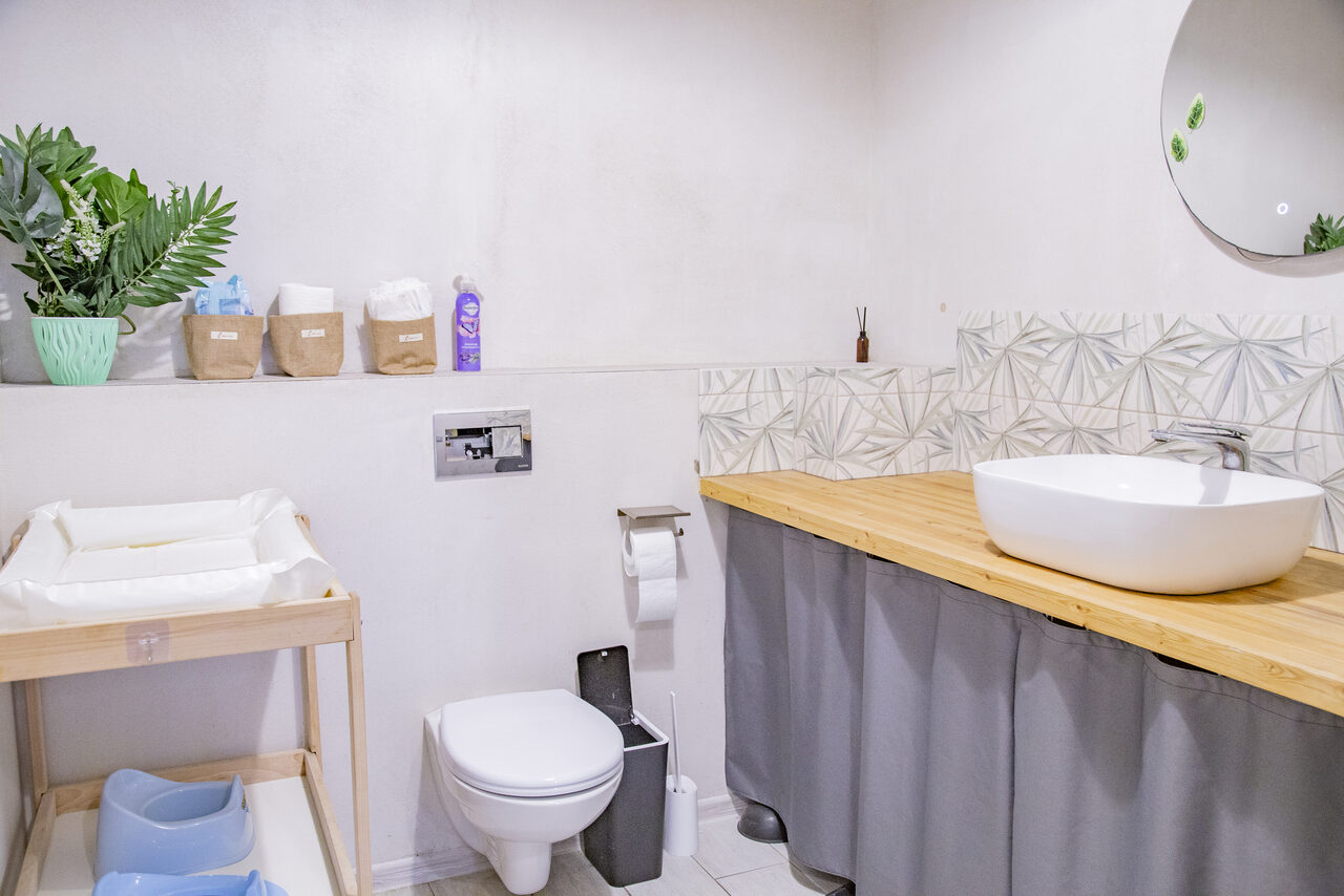 Арендовать Лофт Облака. Зал Баунти на Полярной. Светлая комната с умывальником на деревянной столешнице, круглым зеркалом, пеленальным столиком, унитазом и рулоном туалетной бумаги. Зеленые растения в горшке, контейнеры с аксессуарами и косметикой на полке. Светлые стены, серые декоративные панели.