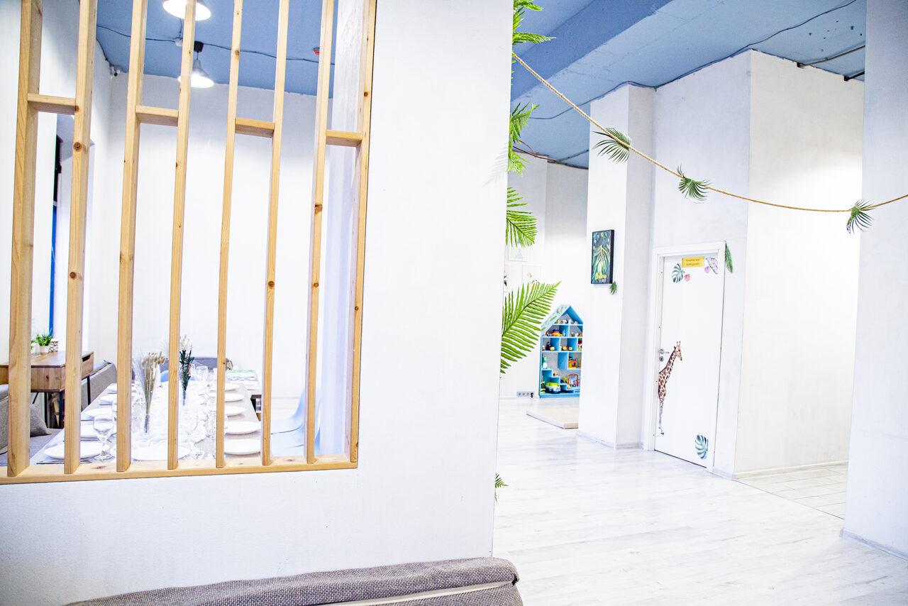 Арендовать Лофт Облака. Зал Баунти на Полярной. Современное пространство с белыми стенами и светлым полом. Деревянная решетка разделяет зоны. Потолок синие, есть подвесные светильники. Видны декоративные элементы в виде зелёных растений и рисунки детской тематики на стенах и двери.