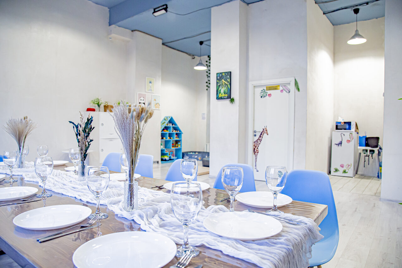 Арендовать Лофт Облака. Зал Баунти на Полярной. Просторное помещение с деревянным столом, расположенным параллельно камере, вокруг голубые стулья. На столе белые тарелки и бокалы. Яркое освещение, светлые стены, в углу разноцветная детская мебель. На стенах картины и украшения.