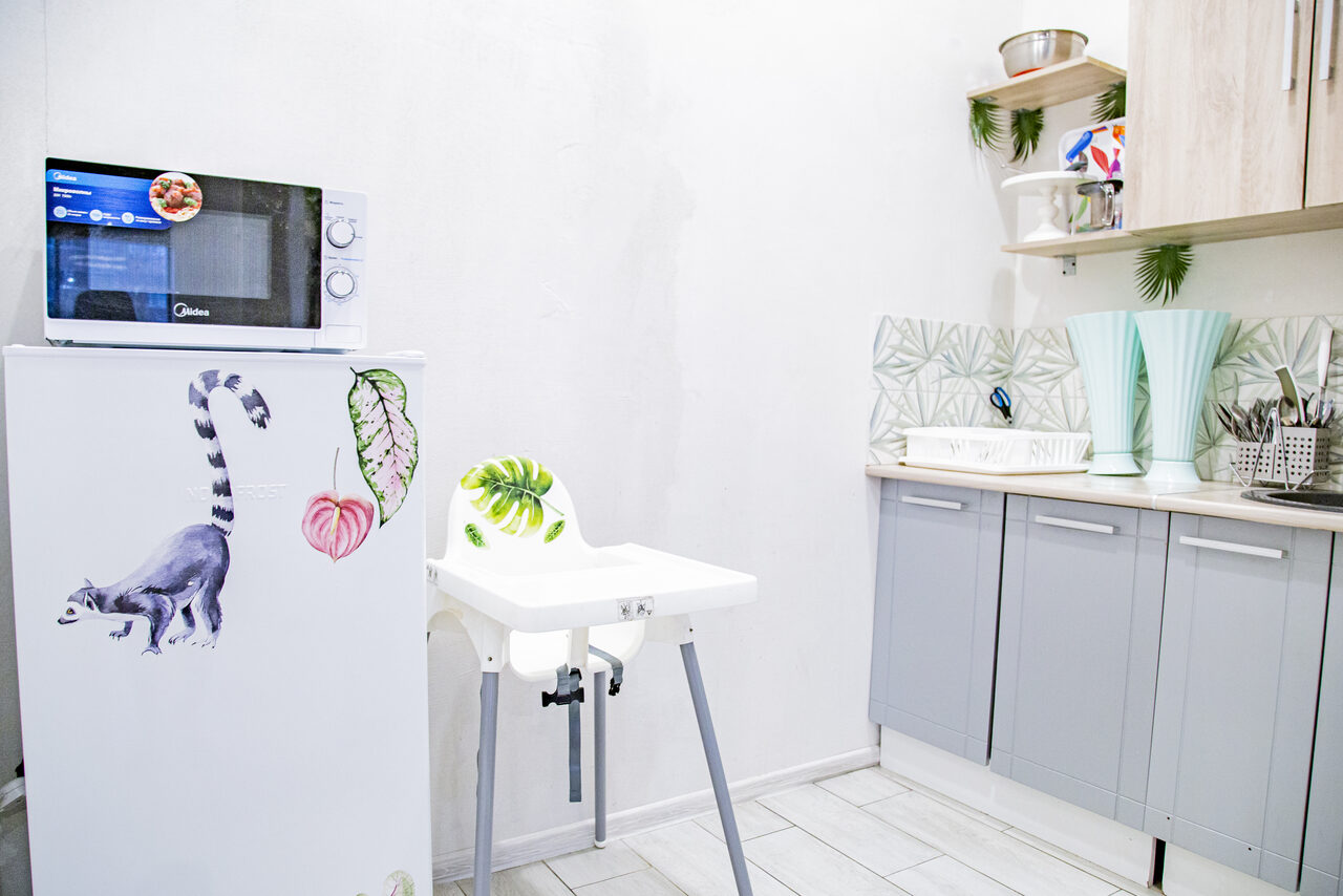Арендовать Лофт Облака. Зал Баунти на Полярной. Кухонное пространство с белым холодильником, украшенным рисунками животных и растений, на котором стоит микроволновка. Рядом детский стульчик. Слева серые шкафчики и открытые полки с посудой. Навесные панели с листовым узором. Светлая плитка на полу.