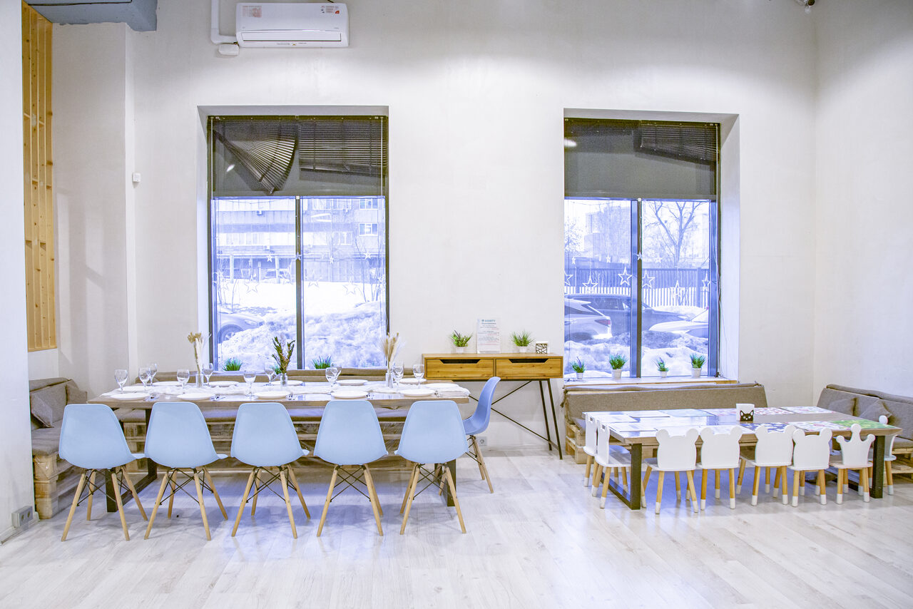 Арендовать Лофт Облака. Зал Баунти на Полярной. Большое помещение с деревянными полами. Справа два больших окна. Слева длинный деревянный стол с голубыми стульями, справа маленький стол с детскими стульями. На стене кондиционер. Между окнами деревянный столик с растениями.