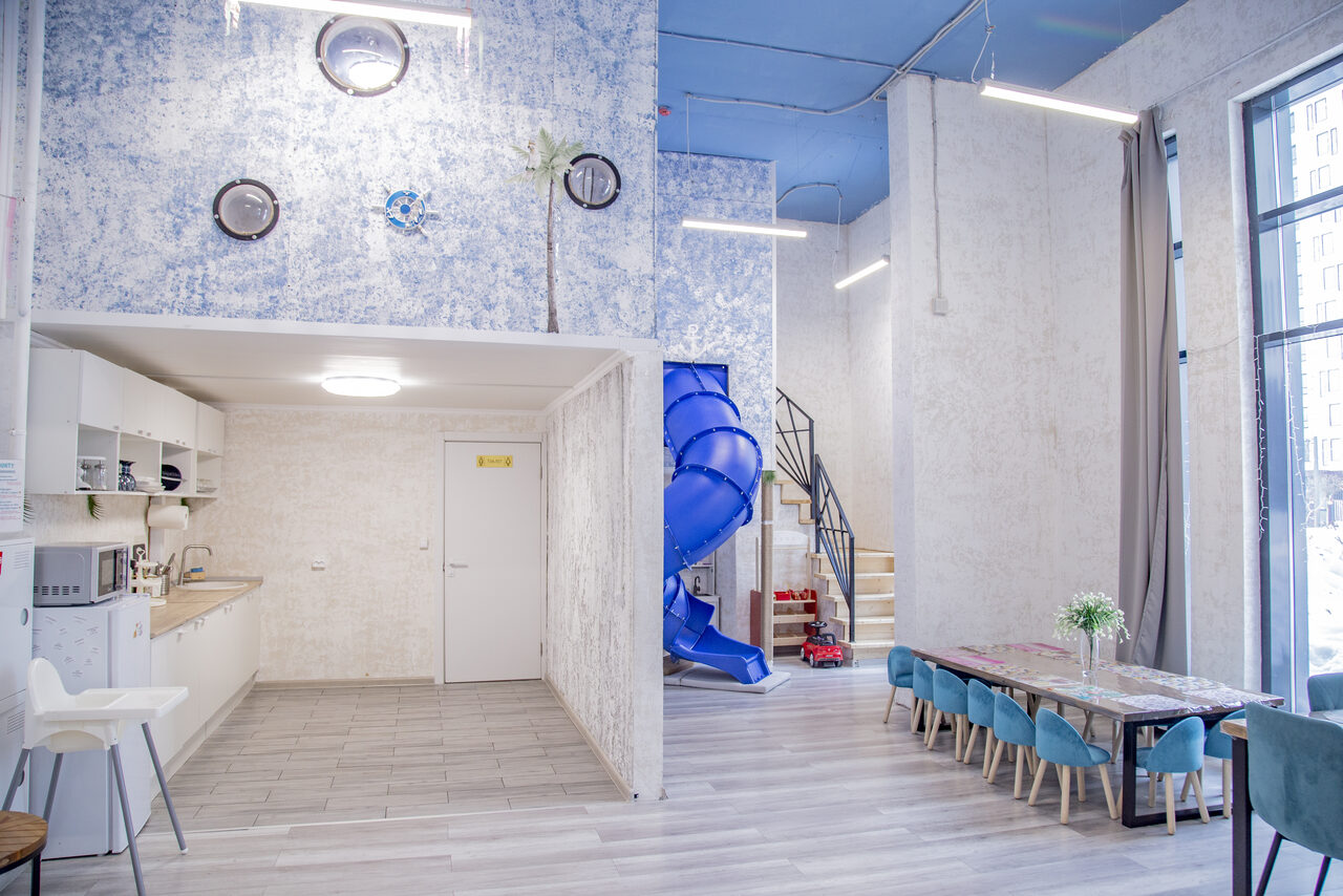 Арендовать Лофт Облака. Зал Баунти на Сельскохозяйственной. Светлое просторное помещение с высокими потолками, кухней слева, серыми шкафами и техникой. Справа обеденный стол с голубыми стульями, рядом лестница и синий винтовой спуск. Светлые стены, большие окна, серый ламинат.