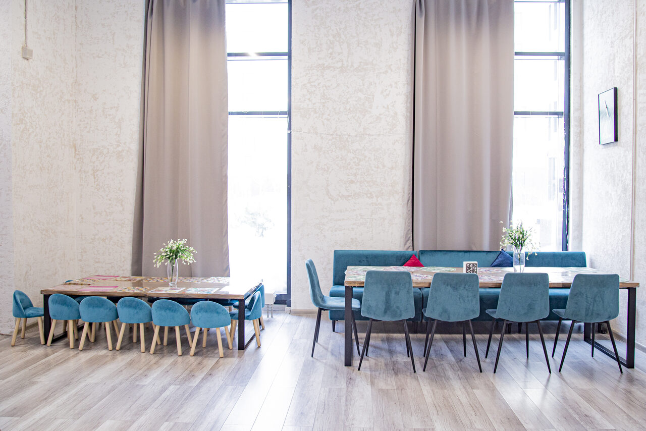 Арендовать Лофт Облака. Зал Баунти на Сельскохозяйственной. Просторное помещение с высокими потолками, двумя большими окнами с бежевыми шторами. Два деревянных стола с сине-зелеными стульями. На столах букеты цветов. Пол светлый деревянный. Стены светло-серые. Справа в верхней части видна рама с картиной.