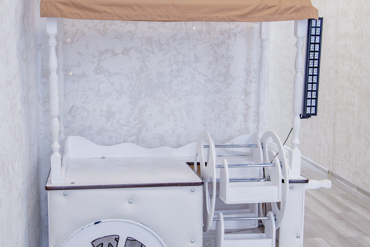 Арендовать Лофт Облака. Зал Баунти на Сельскохозяйственной. Белая деревянная тележка с крышей из светло-коричневой ткани, расположенная у стены с текстурированным белым покрытием. На стене виден узкий вертикальный элемент с квадратными ячейками. Пол светлый, с горизонтальным узором.