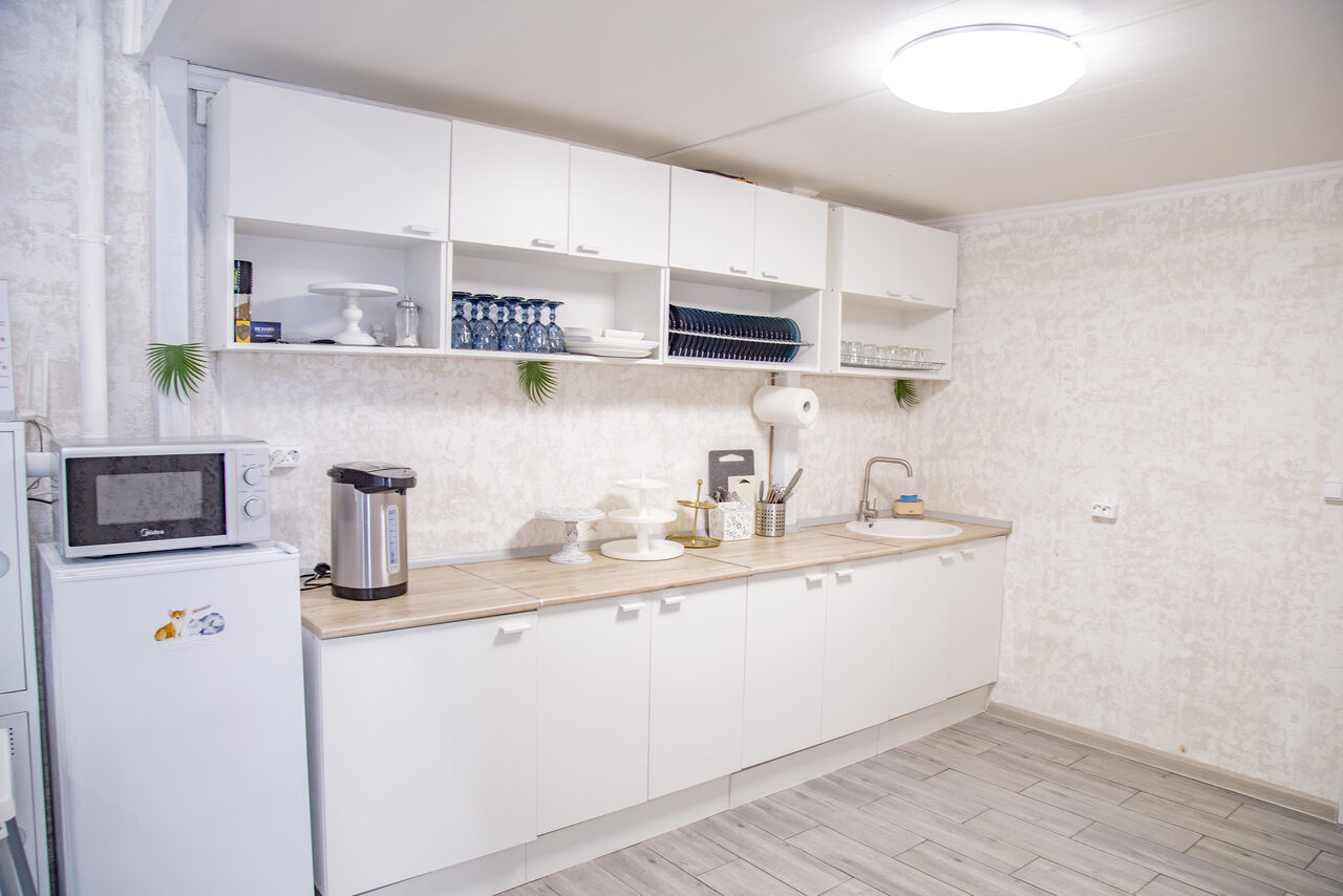 Арендовать Лофт Облака. Зал Баунти на Сельскохозяйственной. Кухонное пространство с длинной рабочей поверхностью, белыми напольными и навесными шкафами. Имеется холодильник, микроволновая печь, электрический чайник и сушилка для посуды. Светлые стены, плиточный пол, круглый потолочный светильник.