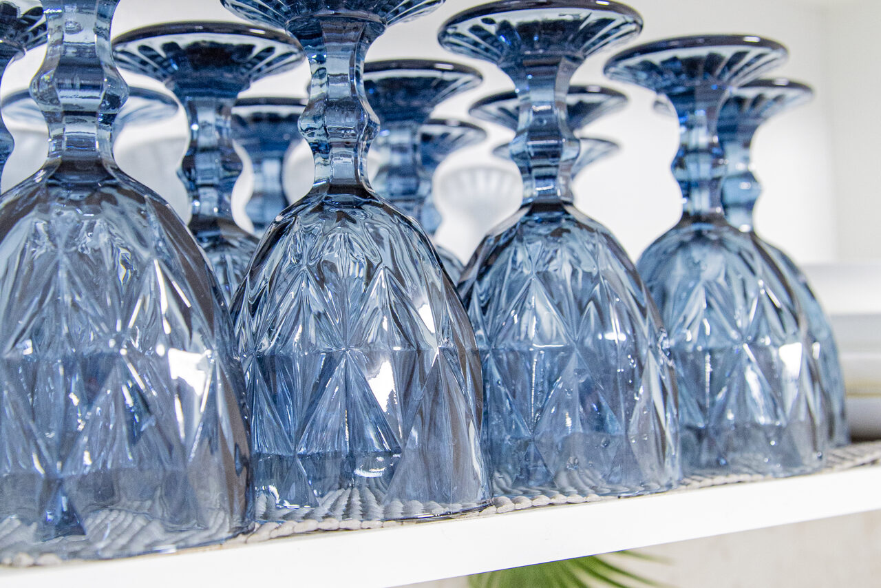 Арендовать Лофт Облака. Зал Баунти на Сельскохозяйственной. Несколько синих стеклянных бокалов перевёрнуты вверх дном на белой полке. Бокалы имеют геометрический узор. Поверхность полки выглядит текстурированной. Видна часть зелёного листа внизу.