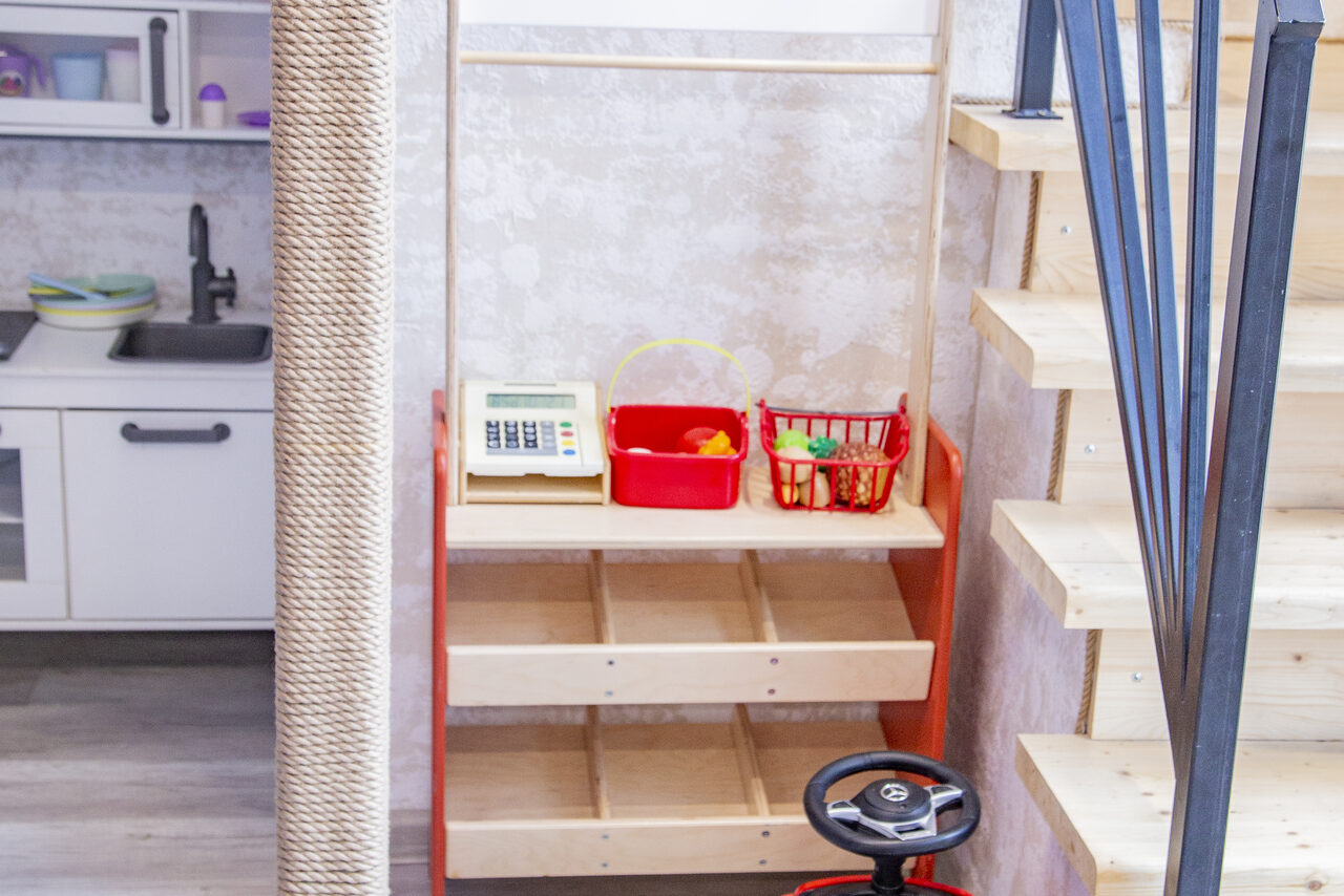 Арендовать Лофт Облака. Зал Баунти на Сельскохозяйственной. Детский уголок со стендом и игрушками, рядом деревянная лестница с металлическими перилами. На заднем плане часть кухни с белыми шкафами и черной мойкой. Освещение естественное. Материалы: дерево и металл. Цвета: светлые, пастельные.