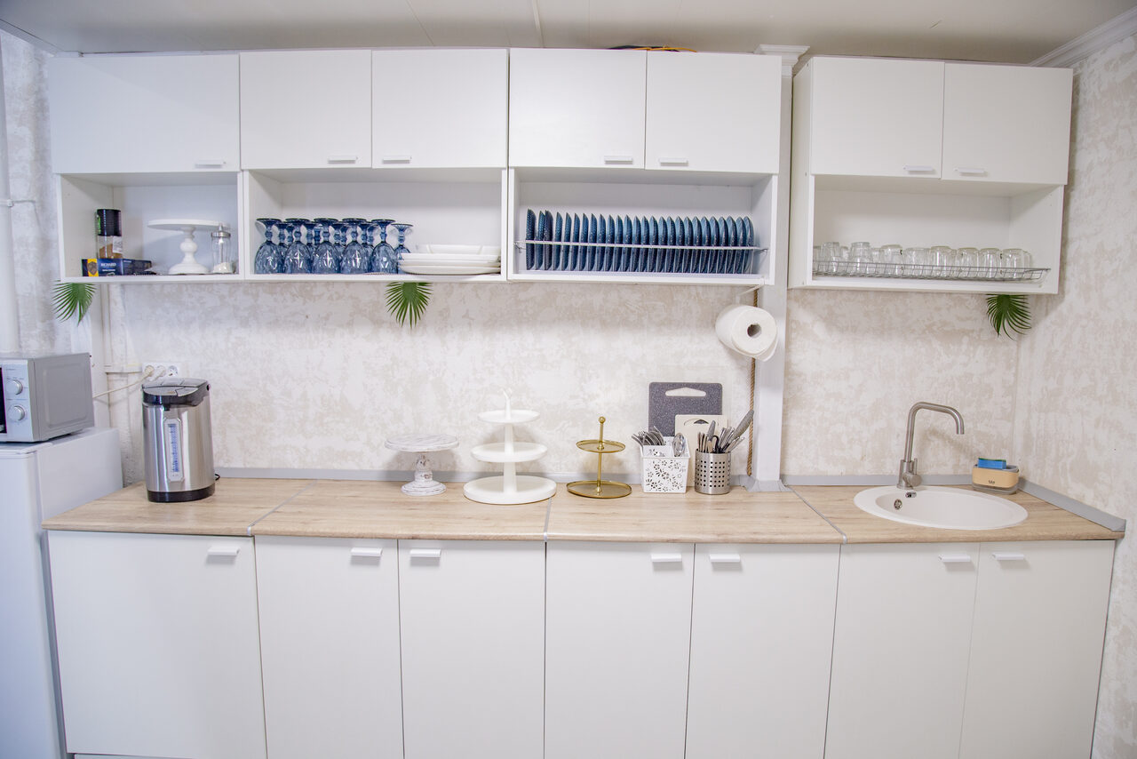 Арендовать Лофт Облака. Зал Баунти на Сельскохозяйственной. Белая кухня с прямой планировкой. Верхние шкафы с распашными дверцами содержат посуду и бокалы. На столешнице металлический чайник, подставка для бумаги, аксессуары и раковина. Светлые стены с текстурой.