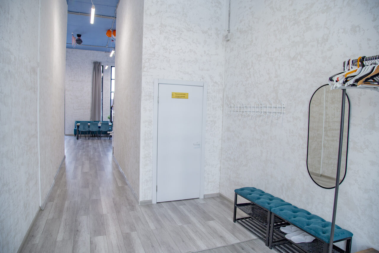 Арендовать Лофт Облака. Зал Баунти на Сельскохозяйственной. Узкий коридор с светлыми стенами и светлым деревянным полом. Металлическая скамья с зелёной обивкой и встроенной полкой. Зеркало овальной формы и стойка с вешалками. Дверь с табличкой, ведущая в просторное помещение с диваном. Потолочное освещение.