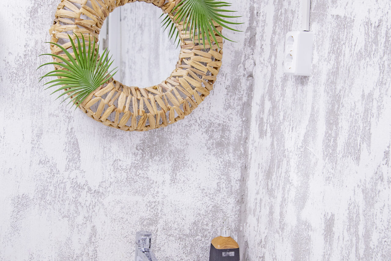 Арендовать Лофт Облака. Зал Баунти на Сельскохозяйственной. Круглое зеркало в деревянной раме с зелеными листьями, серые текстурированные стены, белая розетка, часть смесителя над черной бутылью с мылом.