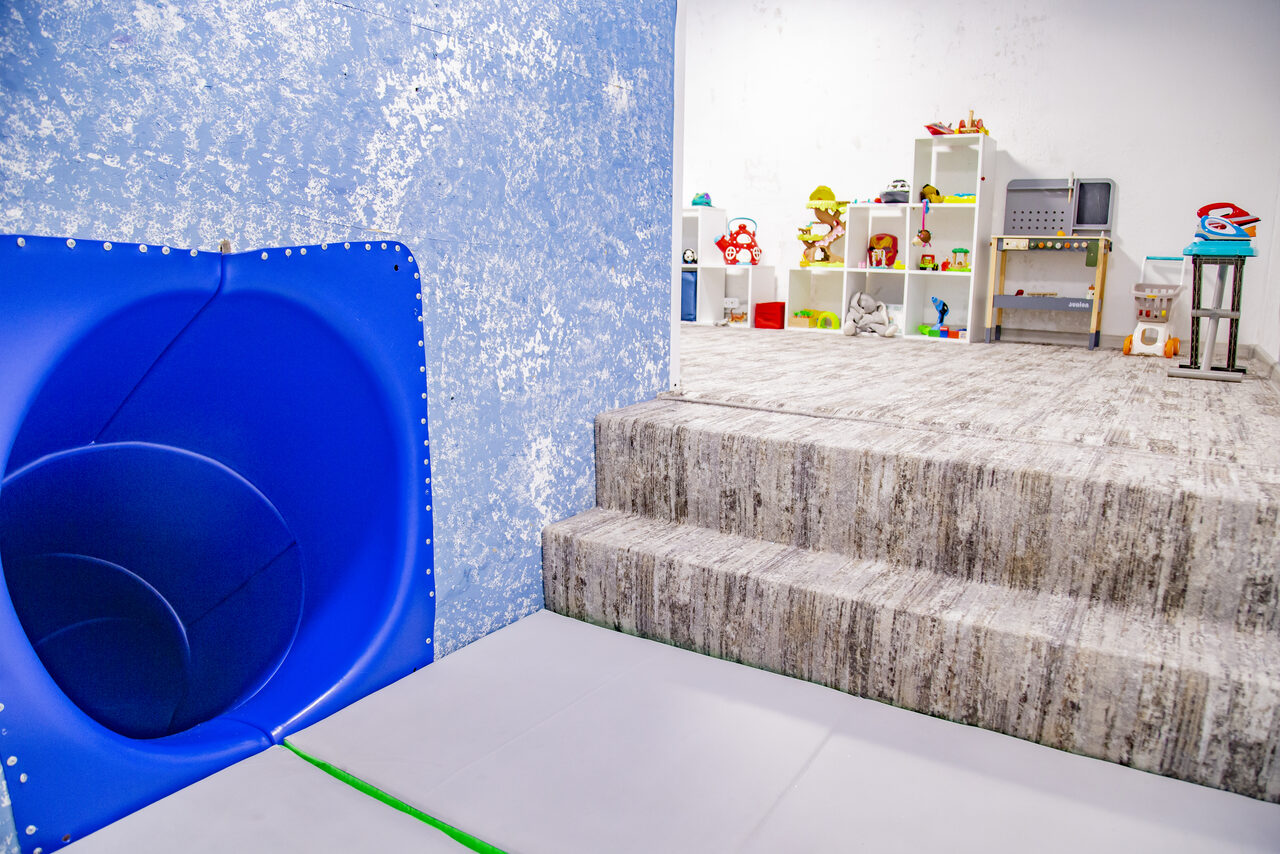 Арендовать Лофт Облака. Зал Баунти на Сельскохозяйственной. Детская игровая комната с голубой горкой, ступеньками, полом с серым ковролином. Игровая зона с белыми стеллажами, игрушками и разноцветными предметами. Светлые стены, естественное освещение.