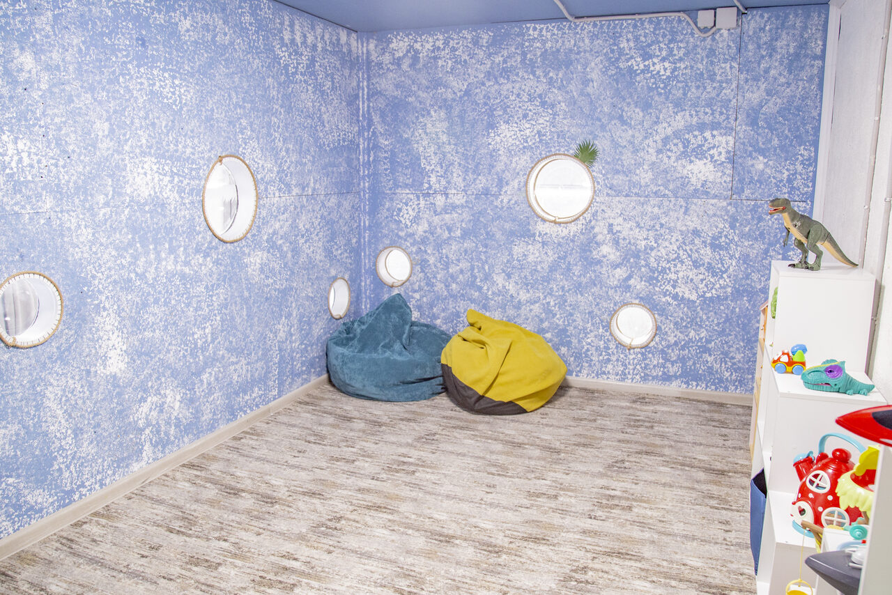 Арендовать Лофт Облака. Зал Баунти на Сельскохозяйственной. Комната с голубыми стенами и круглыми окнами, пол деревянный светлый. Два кресла-мешка, синий и желтый. Белый стеллаж с игрушками, на нем динозавр. В стене вмонтированы круглые зеркала. Освещение яркое.